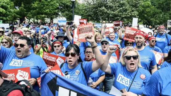 Crowd of educators wearing blue shirts raise their hands in solidarity at a May 1 day of action event in Washington, DC